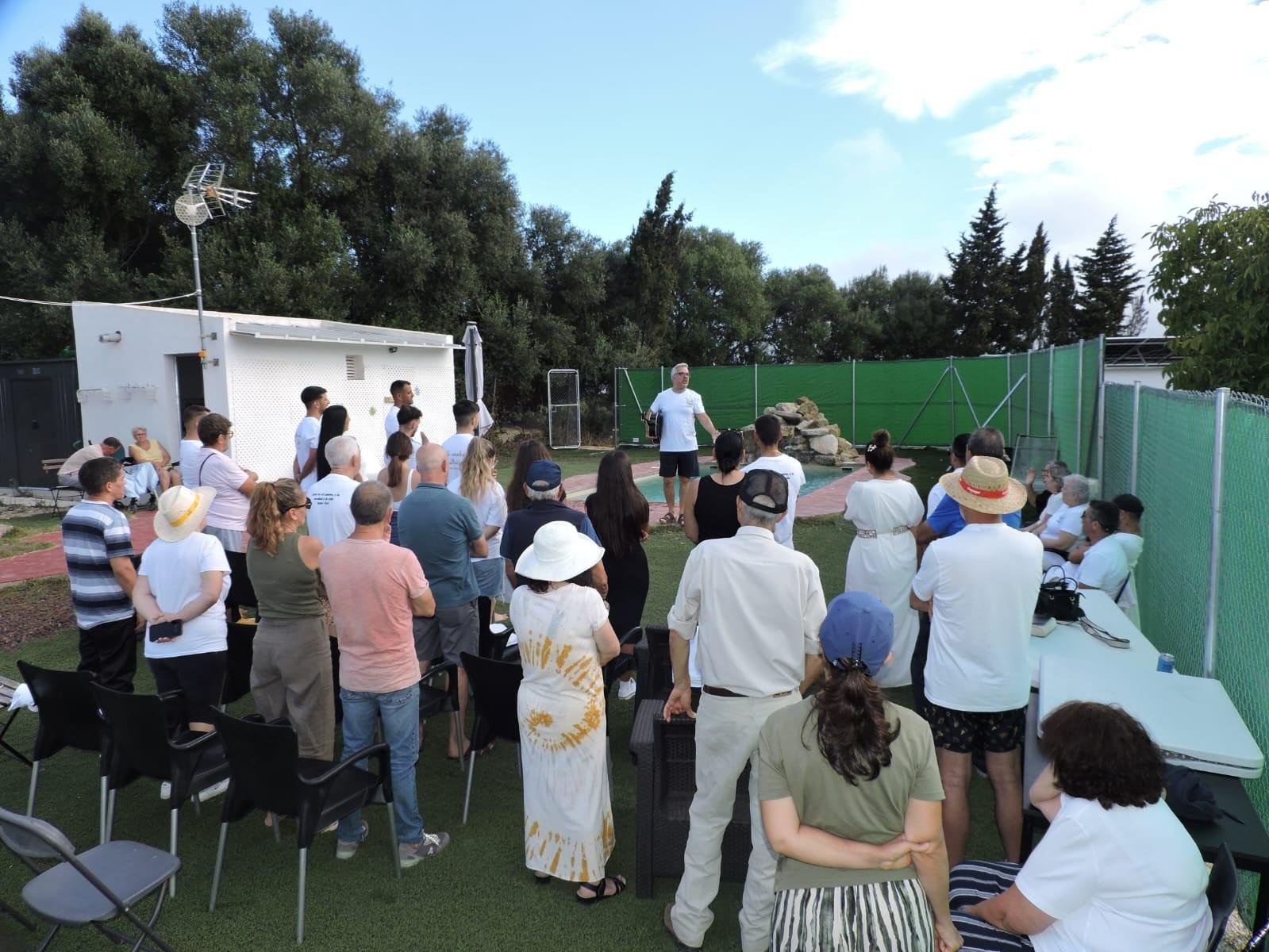 Grupo de la iglesia reunido en un jardín junto a una piscina, escuchando de pie una enseñanza al aire libre