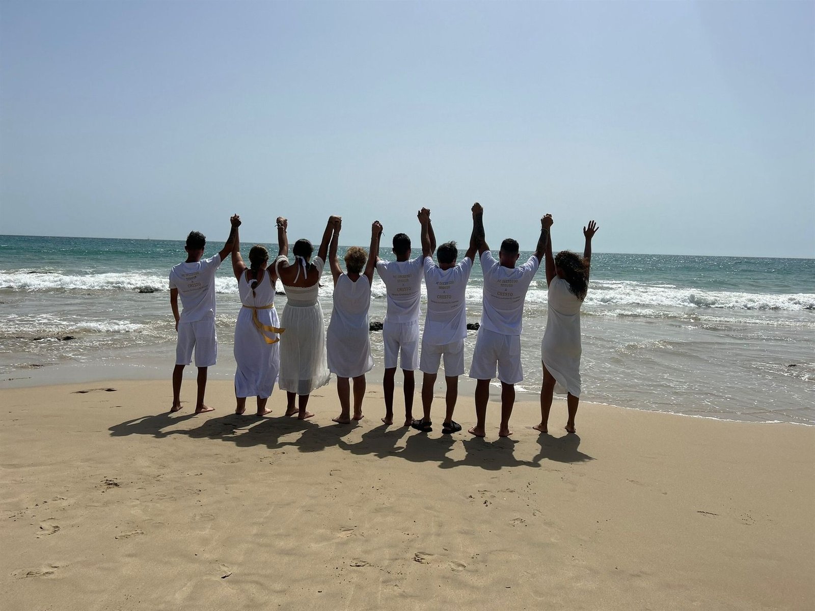 El grupo de la iglesia de espaldas en la playa, cogidos de la mano y con los brazos levantados hacia el mar, en el momento del bautismo